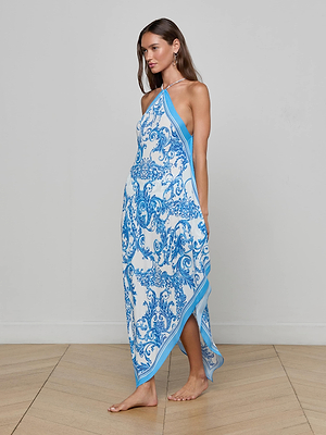 A woman stands barefoot on wooden flooring, wearing the Elise Cover-Up Dress—a blue baroque print halter midi made from water-safe crepe—set against a plain light gray wall.