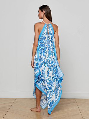 A woman stands barefoot on a wooden floor, facing away. She wears the Elise Cover-Up Dress—a blue baroque print halter midi with an asymmetric hem, crafted from water-safe crepe. Her straight hair falls down her back.