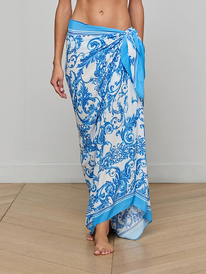 A person wears the Maribel Cover-Up Pareo in blue, tied at the waist, barefoot on a wooden floor against a plain light wall, capturing vintage glamour.