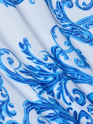 A close-up of the Maribel Cover-Up Pareo shows smooth white fabric with intricate blue floral and swirl patterns, creating an elegant baroque-inspired look. The material appears soft and delicately folded.