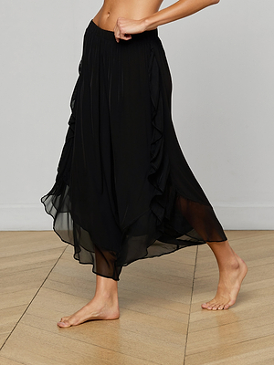 A person stands barefoot on a wooden floor, wearing the Adonis Cover-Up Pant—a flowing black sheer pant with ruffled sides. The image is cropped above the waist, and their left leg is slightly lifted to show the delicate fabric.