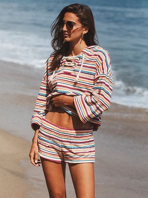 Wearing the Amiya Cover-Up Short and a matching striped sweatshirt, a woman in sunglasses stands on a sandy beach by the ocean, looking away from the camera.