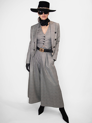 A person wearing a grey three-piece suit with wide-leg trousers, black accessories including the Cebo Belt, and dark sunglasses stands against a white background—a look debuted at New York Fashion Week Fall 2026.