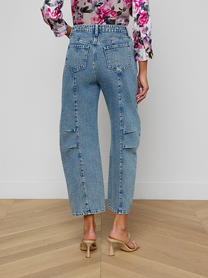 A woman stands indoors on a wooden floor, facing away, wearing Reba Rodeo Jean in denim blue, beige heeled sandals, and a partially visible floral long-sleeve blouse.