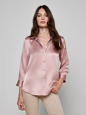A woman with long brown hair wears the Dani Silk Blouse, a shiny light pink button-up, paired with beige pants. She stands against a plain light gray background, looking confidently at the camera.
