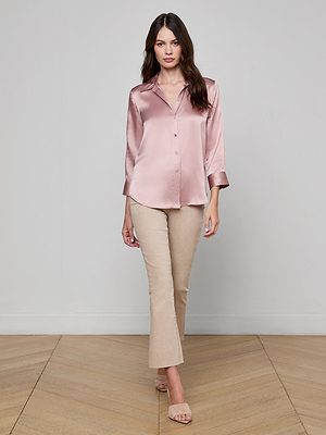 A woman with long brown hair wears the Dani Silk Blouse in pink, paired with beige pants and open-toed sandals, walking indoors on a wooden floor against a plain light-colored wall.