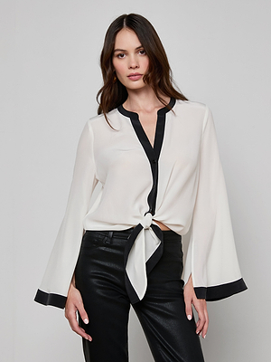 A woman with long, wavy brown hair wears the Charlize Silk Blouse in ivory with black trim and black leather pants, standing against a plain light background.