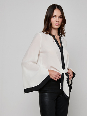 A woman with long brown hair wears the Charlize Silk Blouse in ivory with black trim and flared bell sleeves, paired with black pants. The tie-front waist is cinched as she stands against a plain light background.