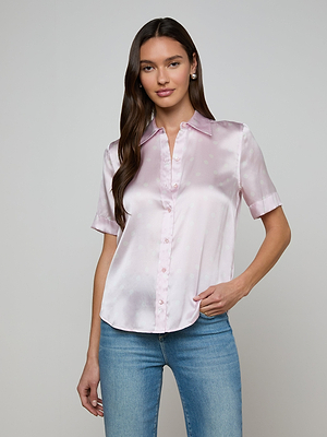 A woman with long brown hair wears the Ellah Silk Blouse in light pink with short sleeves and blue jeans, standing against a plain light gray background.