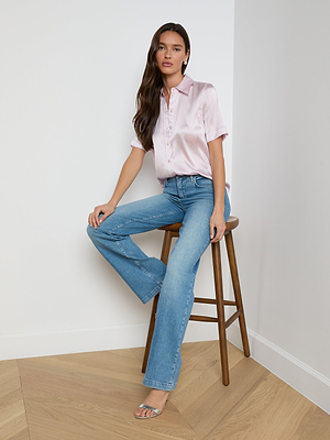 A woman with long, loose hair sits on a wooden stool in a corner, wearing the light pink Ellah Silk Blouse, blue jeans, and clear-strap heels. She looks relaxed. The space has white walls and light wood flooring.