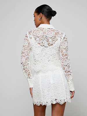 A woman with dark hair in a low bun wears the Roelle Lace Blouse, facing away to showcase its sheer, romantic white floral lace details against a plain light background.
