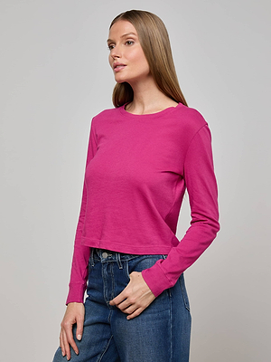 A woman with straight, light brown hair wears the bright pink Zeva Tee and blue jeans, standing with one hand in her pocket and looking off to the side against a plain light gray background.