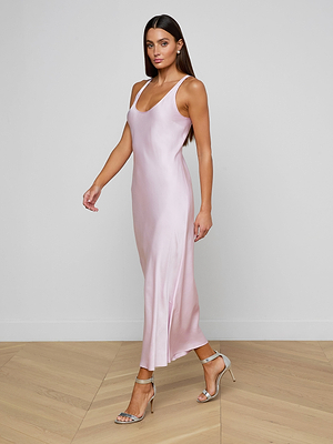 A woman wearing the Akiya Satin Maxi Dress in pastel lilac with silver high-heeled sandals stands on a light wood floor against a plain white wall, walking forward and looking ahead.