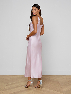A woman in the pastel lilac Akiya Satin Maxi Dress and silver high-heeled sandals poses sideways on a wooden floor, looking over her shoulder against a plain white wall.