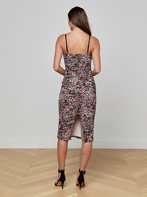 A woman with long brown hair stands facing away, wearing the Caprice Midi Dress in rose cheetah print with thin removable straps and a back slit, paired with black high-heeled sandals on a light wooden floor.