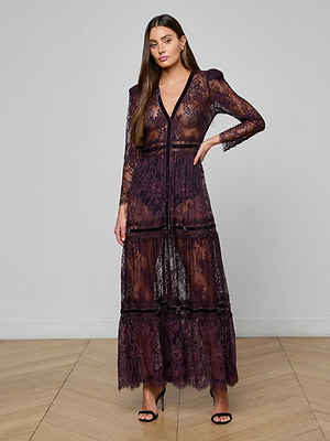 A woman poses on a wooden floor in the Poe Lace Maxi Dress—a long, sheer deep purple gown with velvet trim, long sleeves, and a plunging neckline. She wears black high-heeled sandals; her brown hair falls loose against a plain light gray backdrop.
