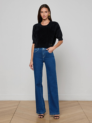 A woman with long brown hair stands indoors on a wooden floor against a plain light gray wall, wearing the L'AGENCE Guinevere Cropped Pullover in black, high-waisted blue jeans, and black open-toe heels.