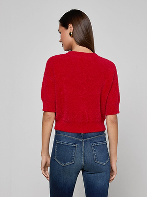 A woman with straight brown hair and round earrings faces away, wearing the Guinevere Cropped Pullover—an ultra-soft, fuzzy red knit crewneck sweater—paired with blue jeans against a light gray background.