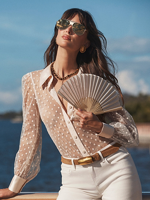 A stylish woman by the water looks up at the sun, wearing sunglasses, white pants, gold accessories, and the Sophie Long-Sleeved Blouse with a white folding fan in hand.