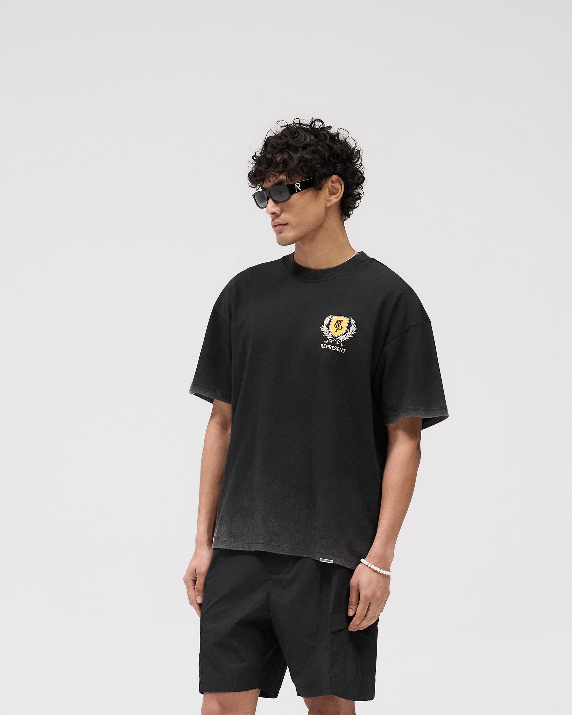 Man wearing a vintage black graphic t-shirt on a plain white background | Crest T-Shirt in Vintage Black | Oversized fit tee with puff and screen print graphics | SS25 T-Shirts | Represent Clo
