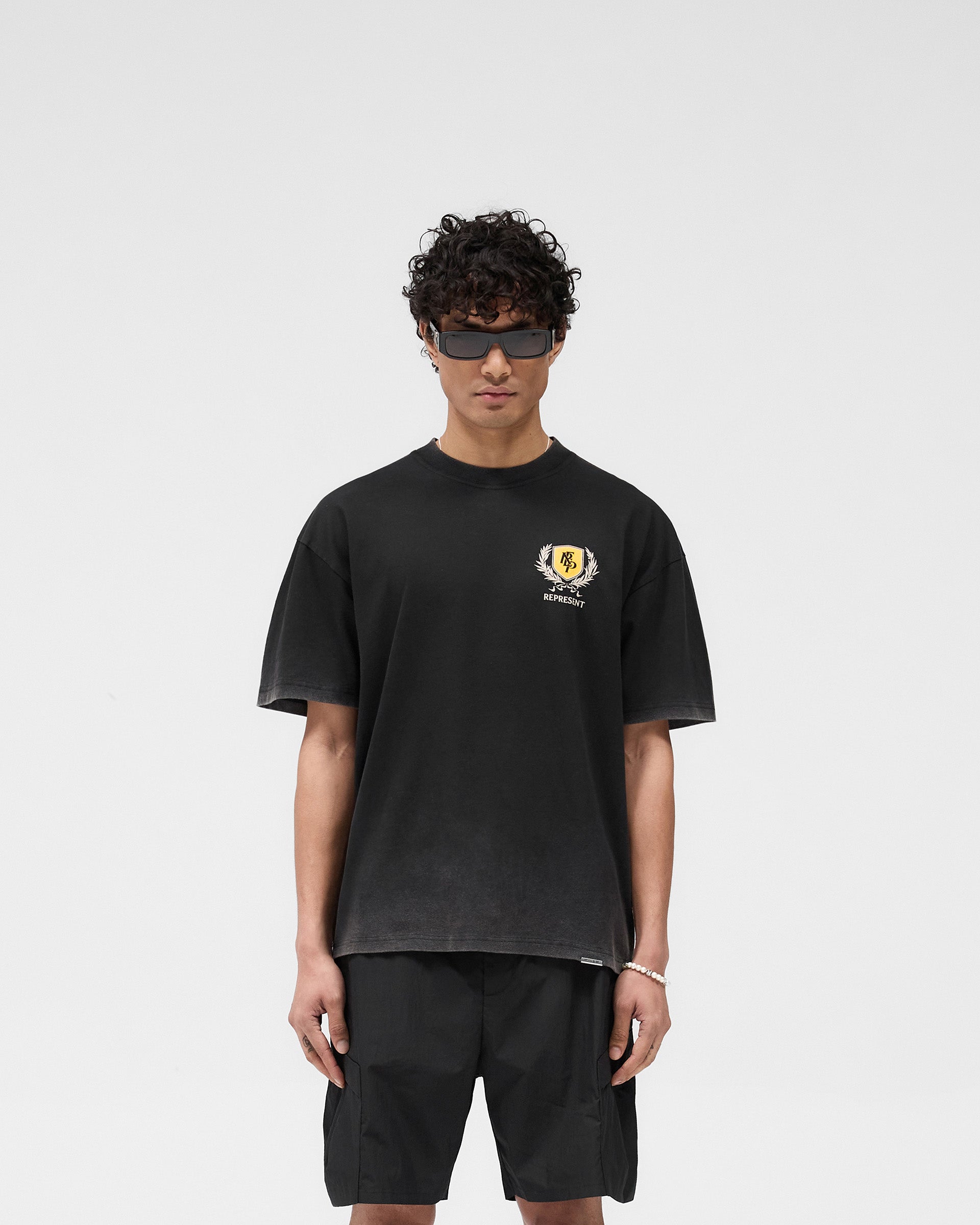 Man wearing a black graphic t-shirt on a plain white background | Crest T-Shirt in Vintage Black | Oversized tee with crest graphic and fade-out wash | SS25 T-Shirts | Represent Clo