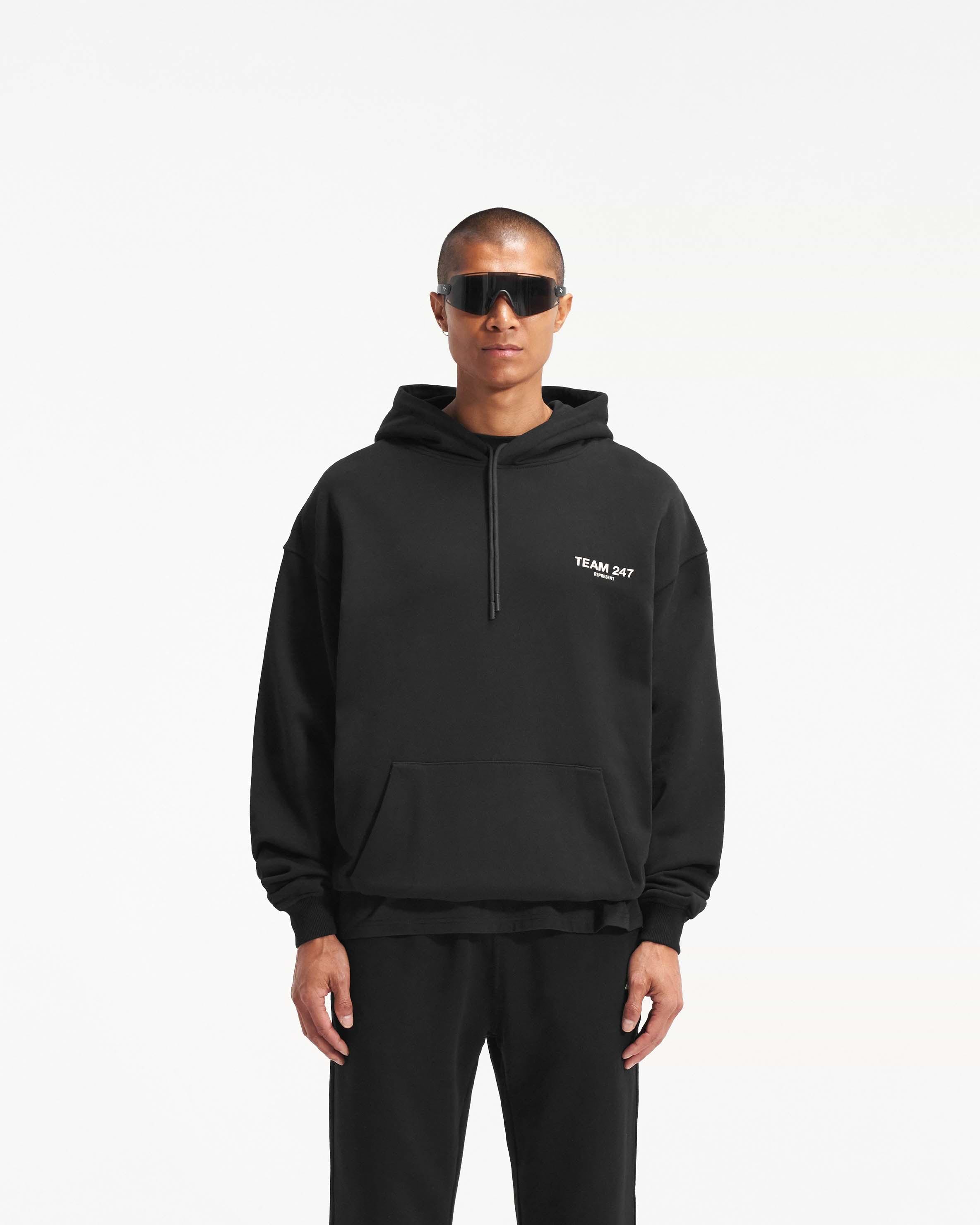 Man wearing a black oversized hoodie on a plain white background | Team 247 Oversized Hoodie in Black | Oversized hoodie with ribbed cuffs and Team 247 print | 247 Hoodies | Represent Clo
