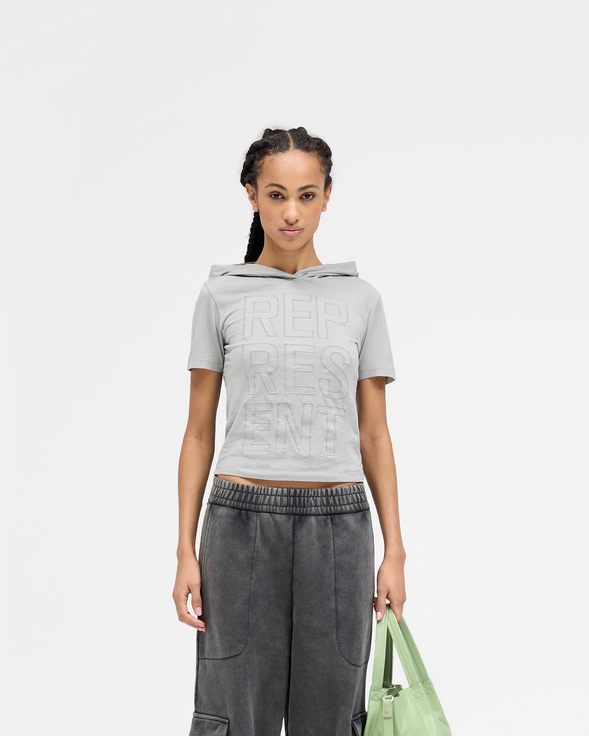Woman wearing a grey t-shirt with hood on a plain white background | Brake Baby T-Shirt in Ultimate Grey | Shrunken fit hooded t-shirt with applique branding | Womens SS25 Tops | Represent Clo
