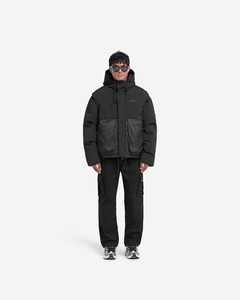 Man wearing a black puffer jacket on a plain white background | Layered Hooded Puffer in Jet Black | Duck down filled puffer with adjustable hood and tonal branding | FW24 Outerwear | Represent Clo