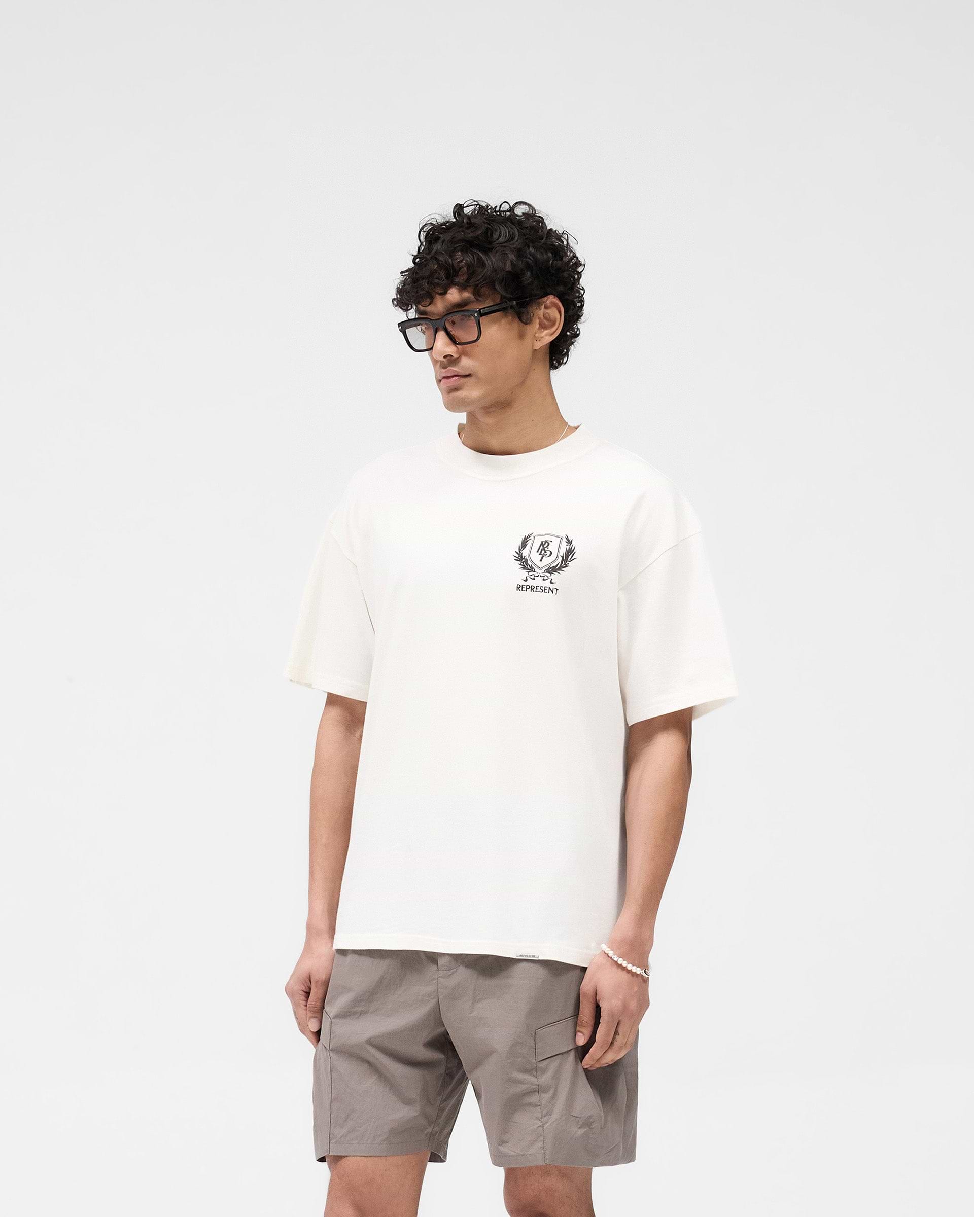 Man wearing a white oversized t-shirt on a plain white background | Crest T-Shirt in Flat White | Premium tee with puff print crest graphics | SS25 T-Shirts | Represent Clo