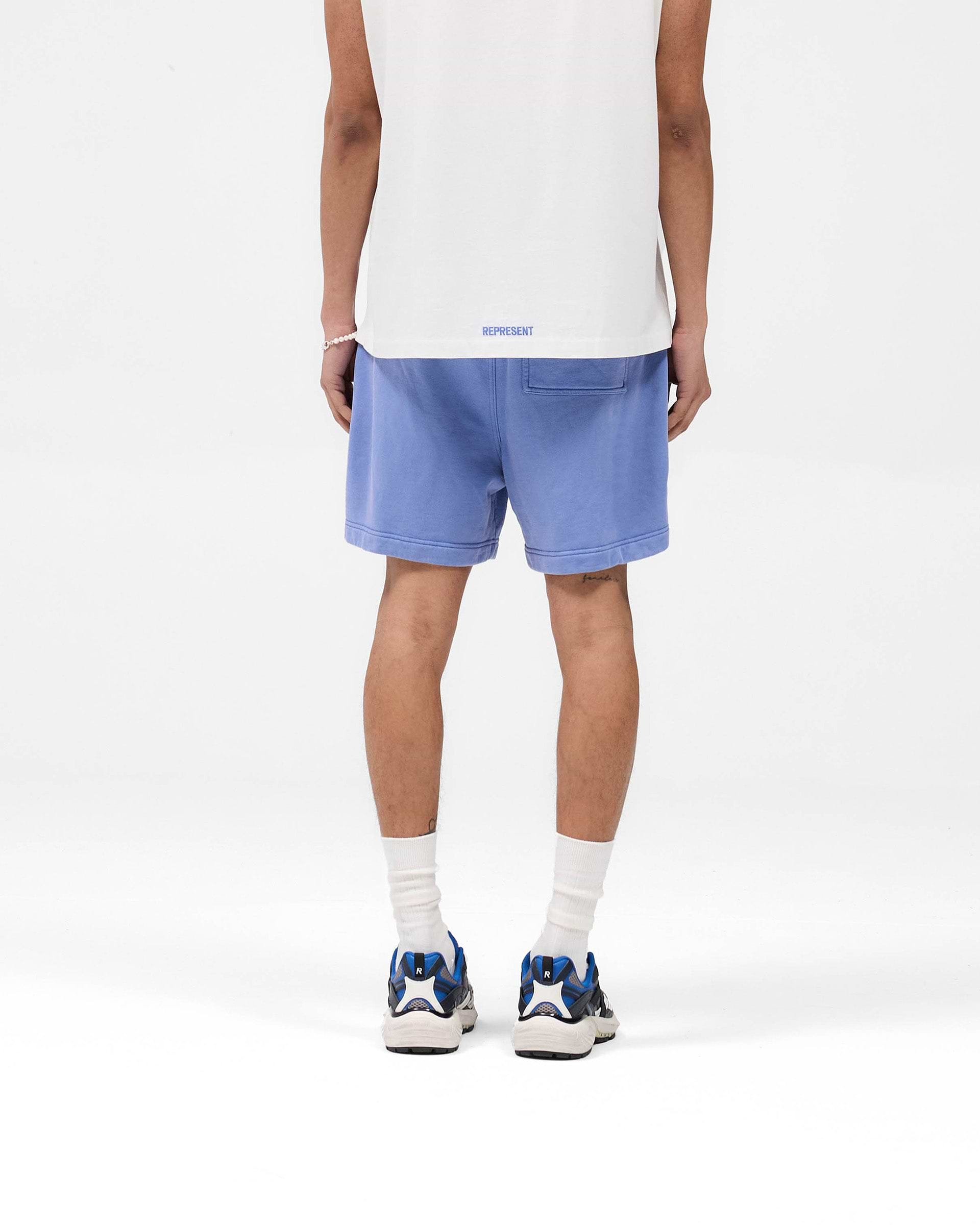Man wearing bright blue graphic shorts on a plain white background | Competition Department Shorts in Bright Blue | 100% cotton shorts with fade-out wash and internal drawcord | SS25 Shorts | Represent Clo