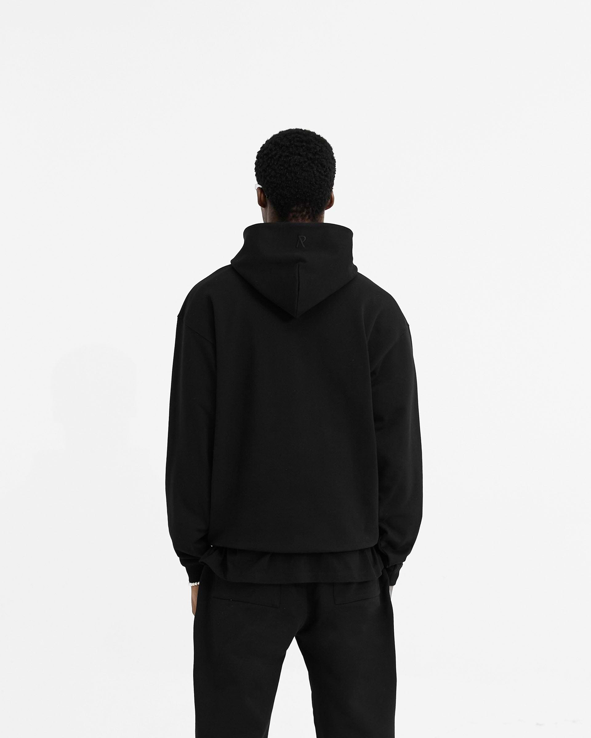 Man wearing a black oversized hoodie on a plain white background | Initial Oversized Hoodie in Black | Heavyweight hoodie with kangaroo pocket and embroidered details | Initial Hoodies | Represent Clo