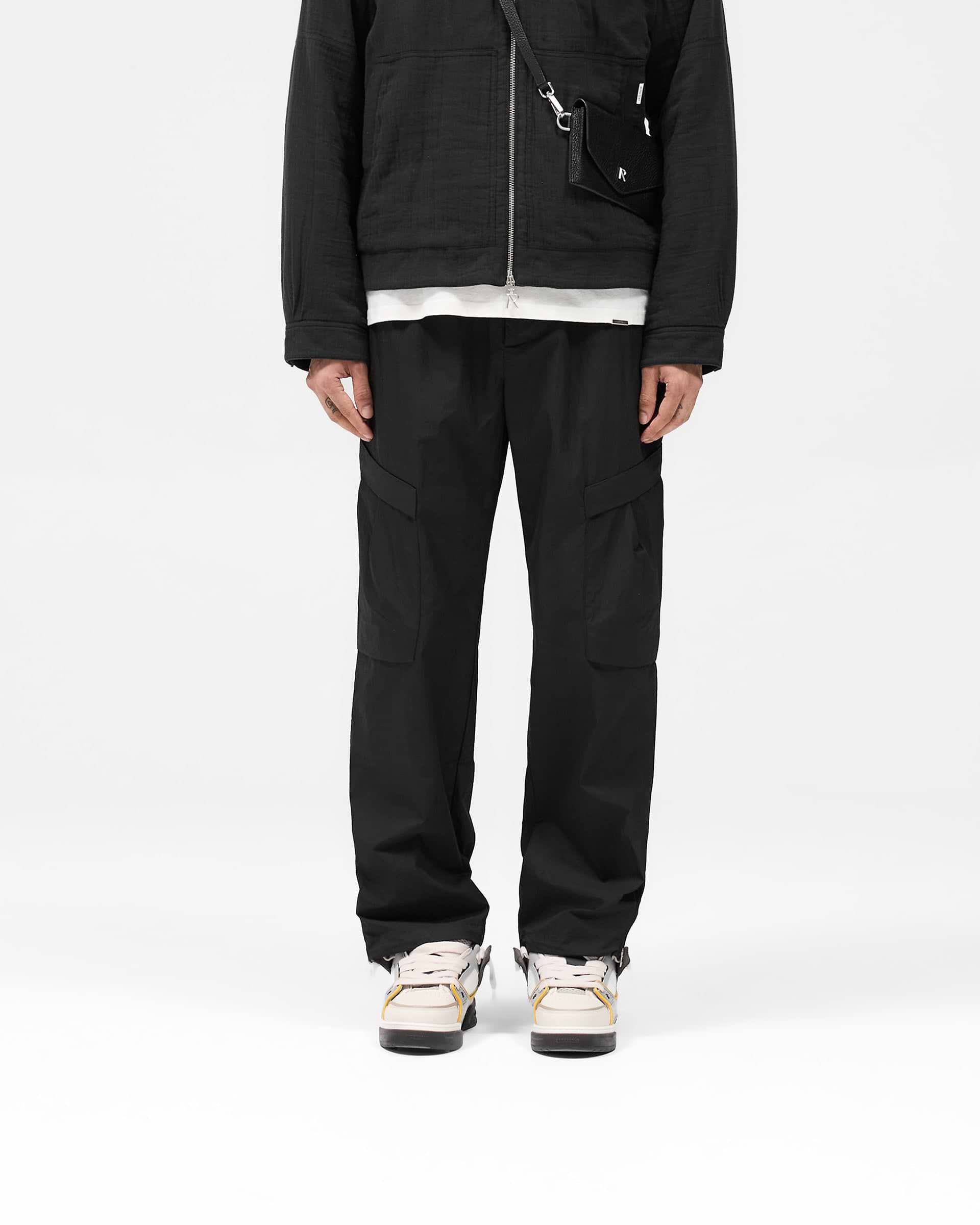 Man wearing black cargo pants on a plain white background | Tech Cargo Pant in Jet Black | Cargo pants with pleated pockets and adjustable hem | SS25 Pants | Represent Clo