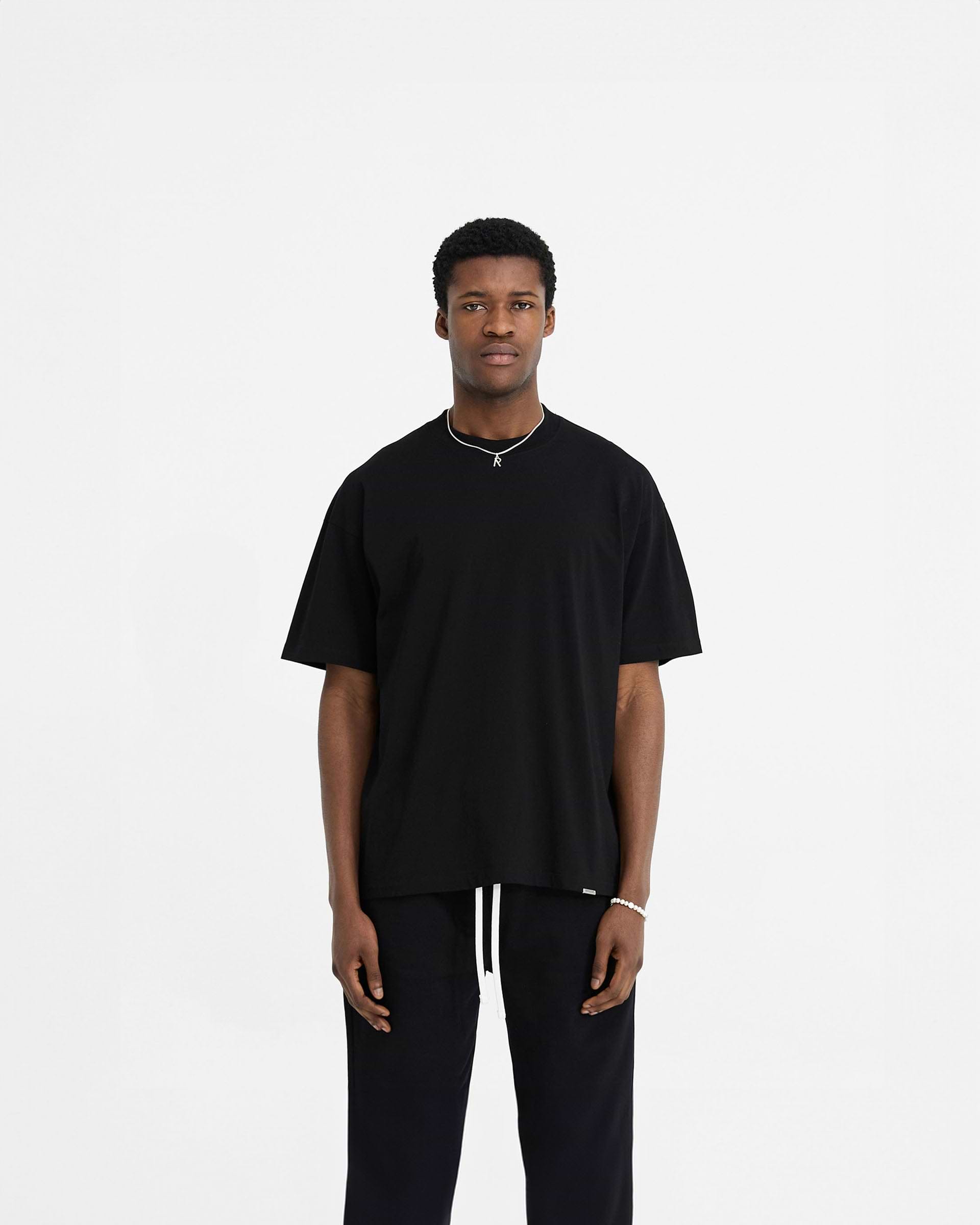 Man wearing a black oversized t-shirt on a plain white background | Initial Oversized T-Shirt in Black | Oversized t-shirt with embroidered details | Initial T-Shirts | Represent Clo