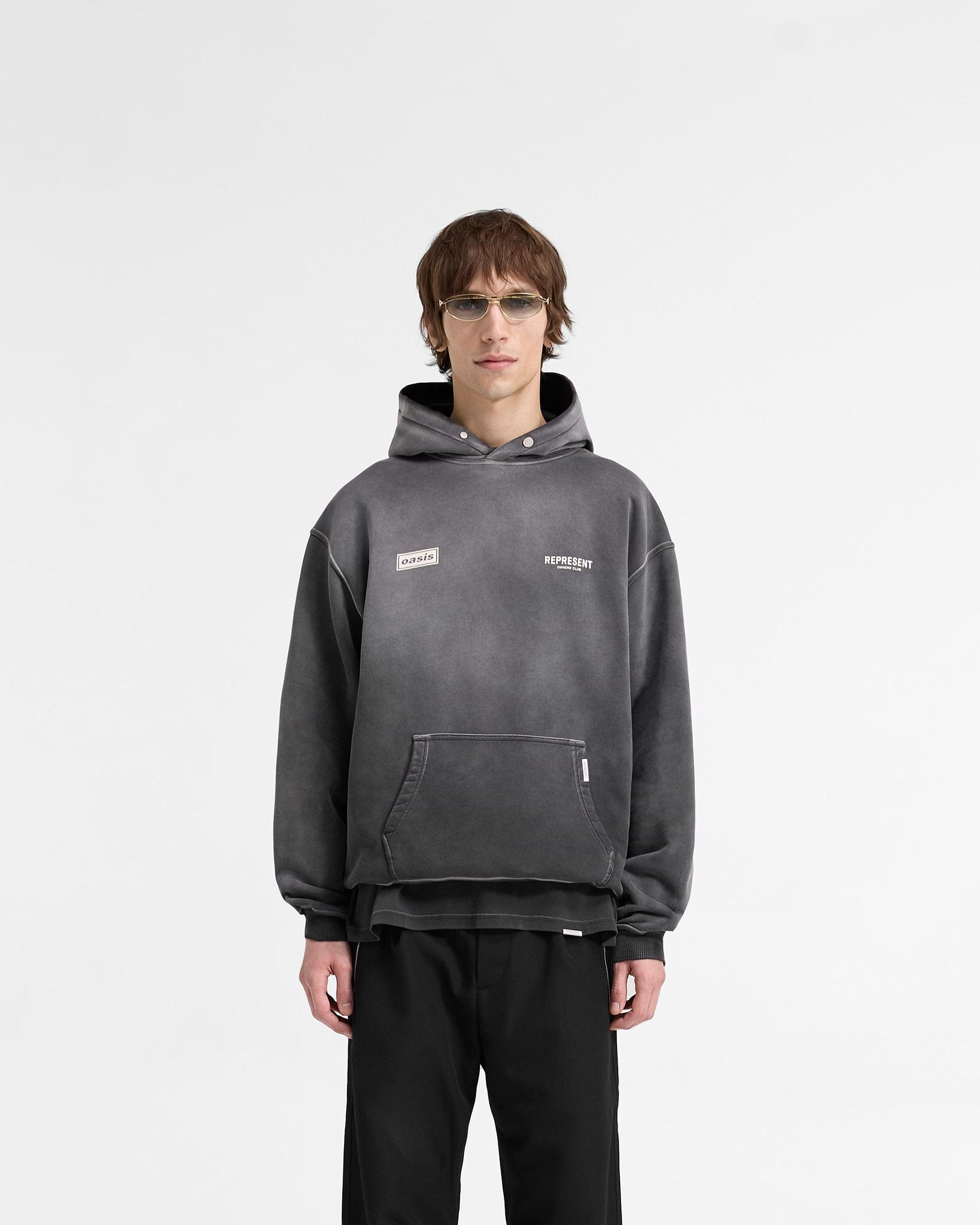 Man wearing an oversized black hoodie with graphic detailing on a plain white background | Represent X Oasis Owners Club Hoodie in Stained Black | Oversized cotton hoodie with exclusive band artwork | FW24 Hoodies | Represent Clo