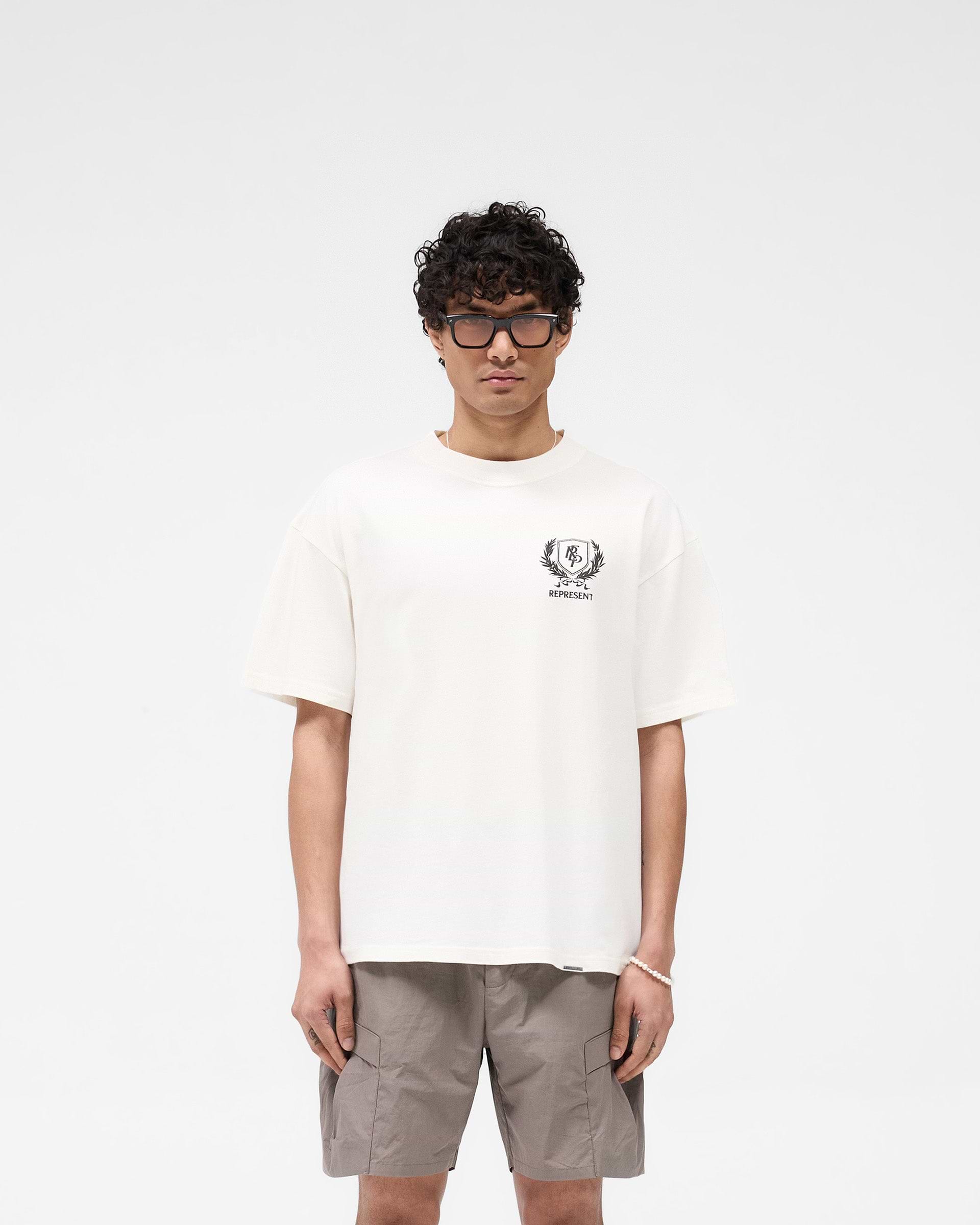 Man wearing a white graphic t-shirt on a plain white background | Crest T-Shirt in Flat White | Oversized tee with crest and Overdrive branding | SS25 T-Shirts | Represent Clo