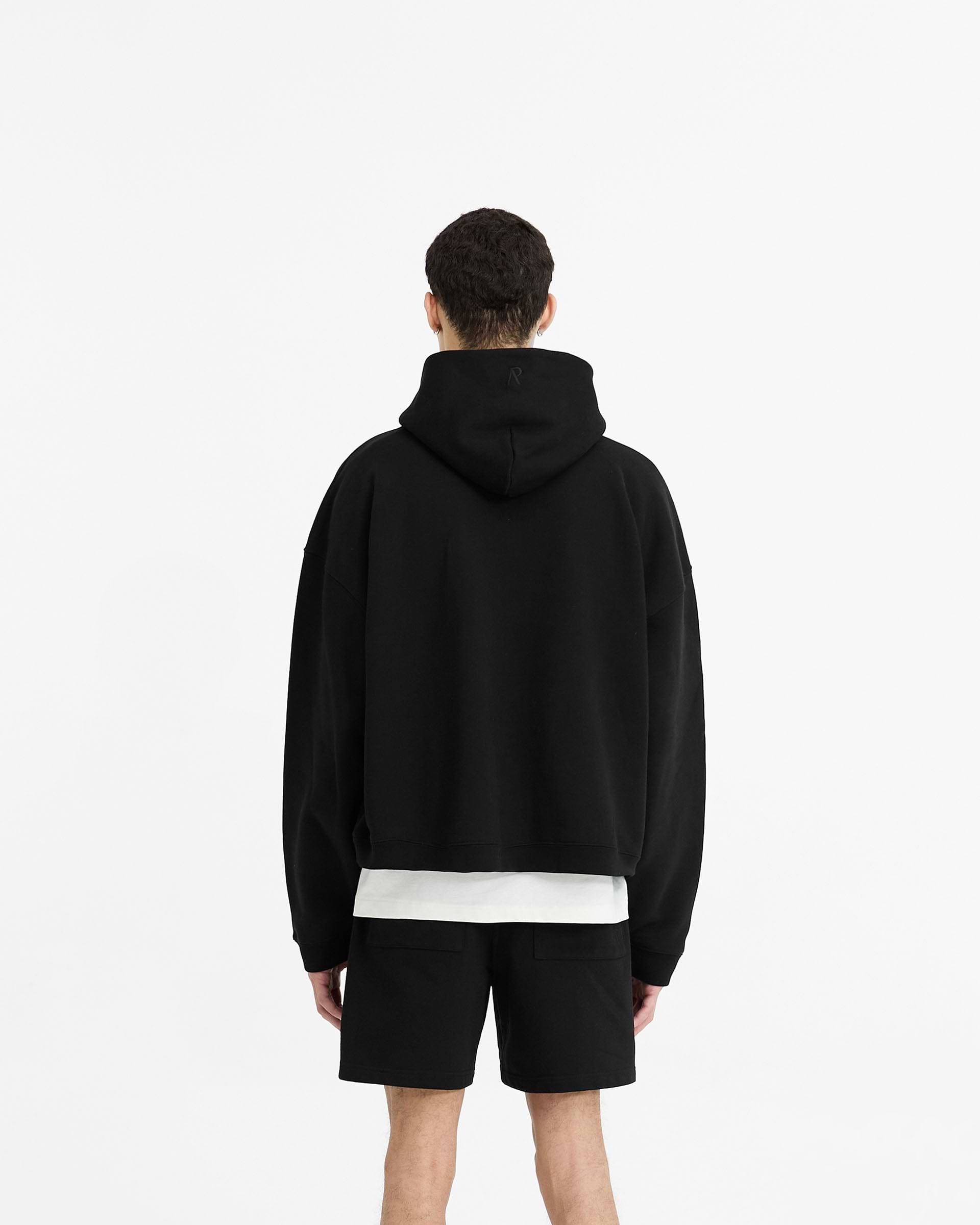 Man wearing an oversized black hoodie on a plain white background | Initial Boxy Hoodie in Black | Boxy cropped hoodie with wide sleeves | Initial Hoodies | Represent Clo