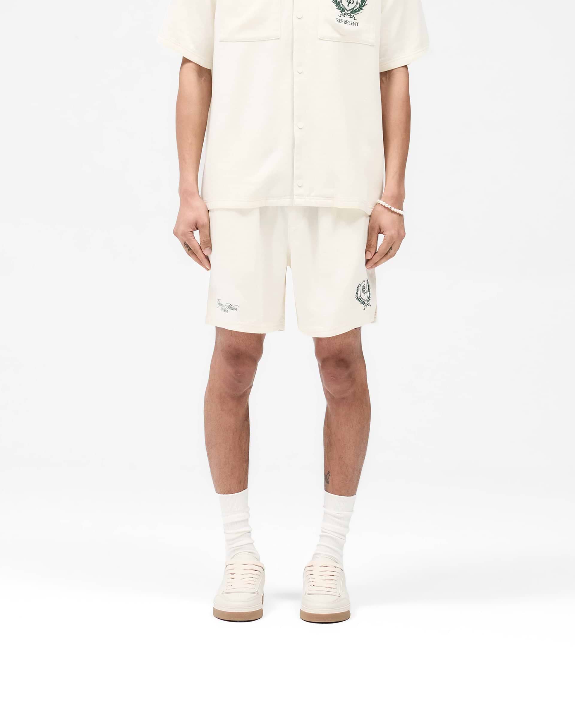 Man wearing antique white shorts on a plain white background | Represent Crest Short in Antique White | Shorts with elasticated waistband and crest embroidery | SS25 Shorts | Represent Clo