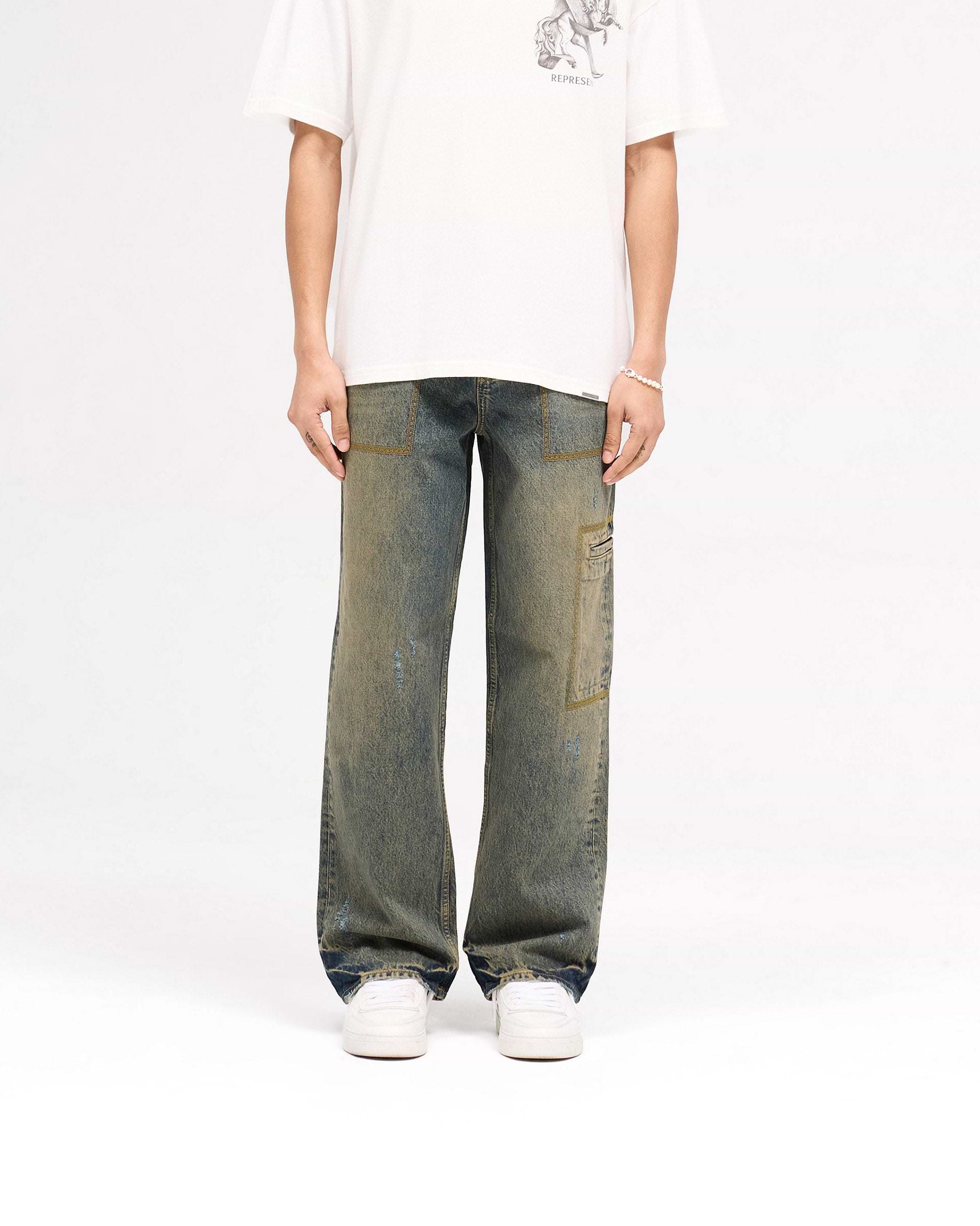 Man wearing blue denim jeans on a plain white background | R3 Drive Denim in Tobacco Blue | Baggy fit denim with distressed detailing | SS25 Denim | Represent Clo