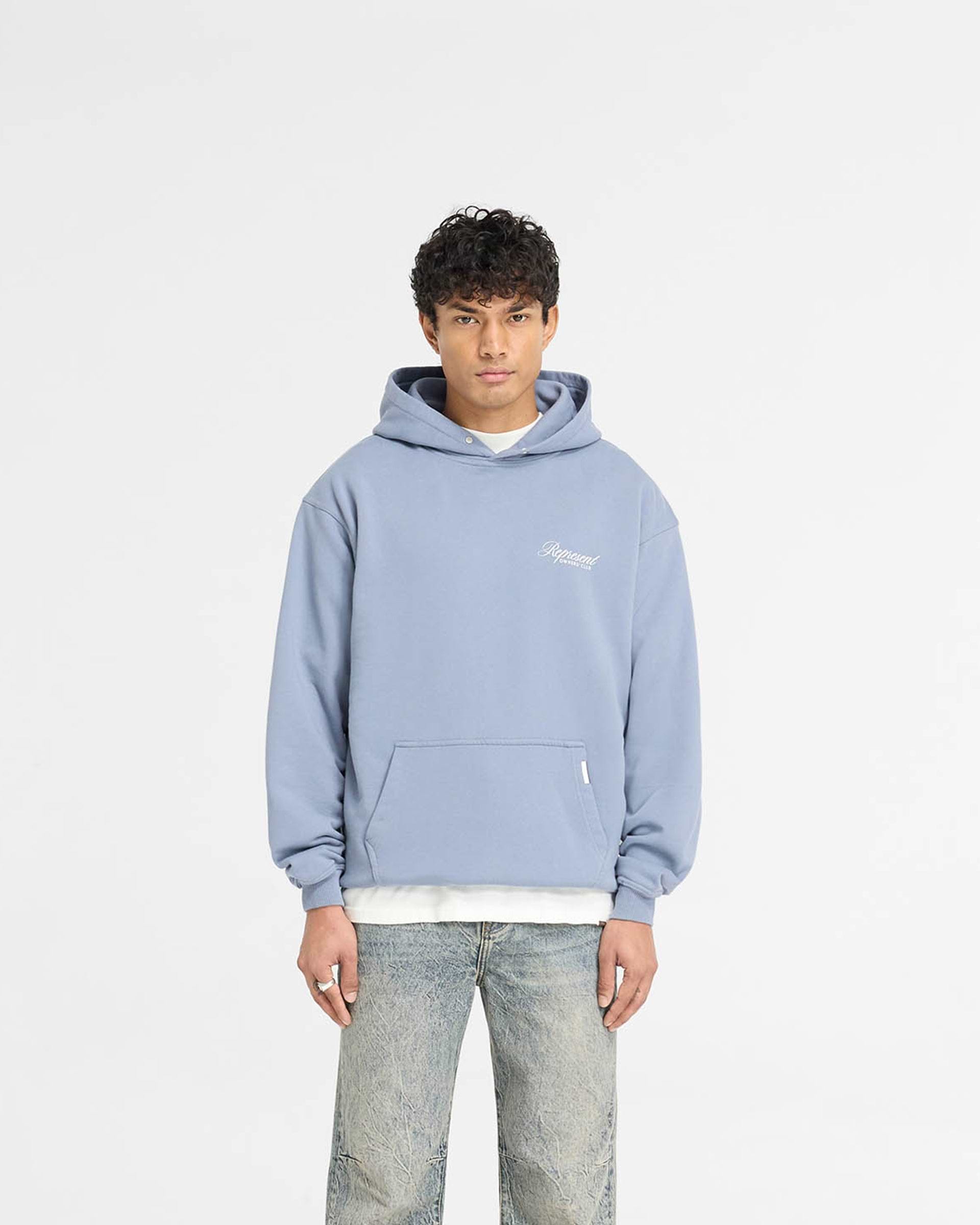 Man wearing a dusty blue hoodie on a plain white background | Represent Owners Club Script Hoodie in Dusty Blue | Oversized fit hoodie with ribbed trims | Owners Club Hoodies | Represent Clo