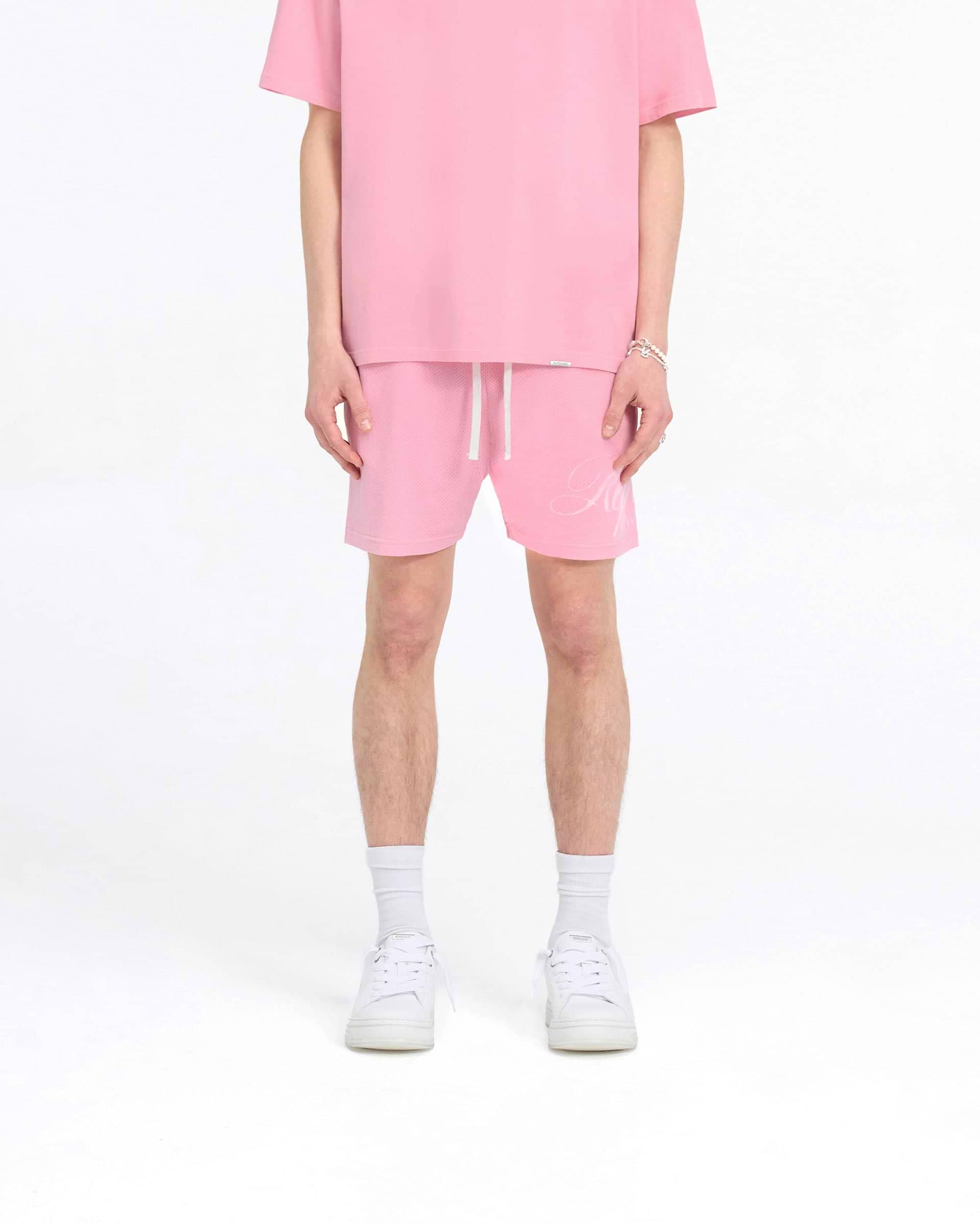 Man wearing pink mesh shorts on a plain white background | Represent Owners Club Script Mesh Shorts in Pink | Mesh shorts with elasticated waistband | Owners Club Shorts | Represent Clo