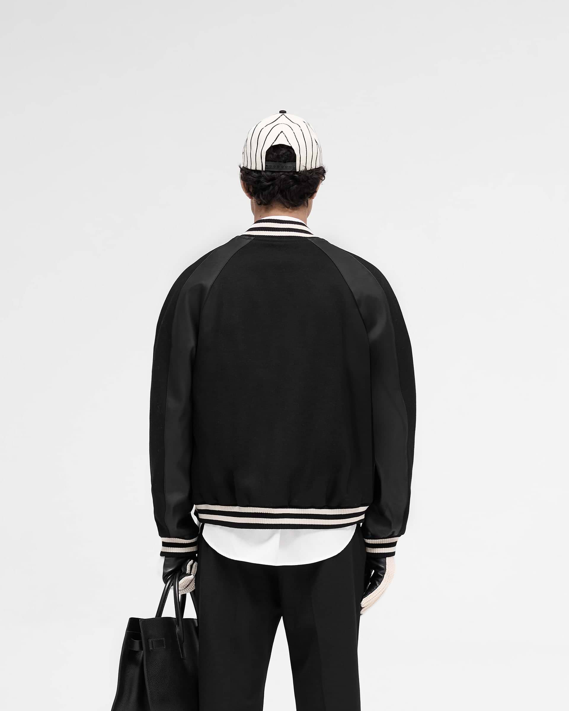 Man wearing a black raglan varsity jacket on a plain white background | Raglan Varsity Jacket in Black | Oversized fit with raglan sleeves and zip pockets | SS25 Outerwear | Represent Clo