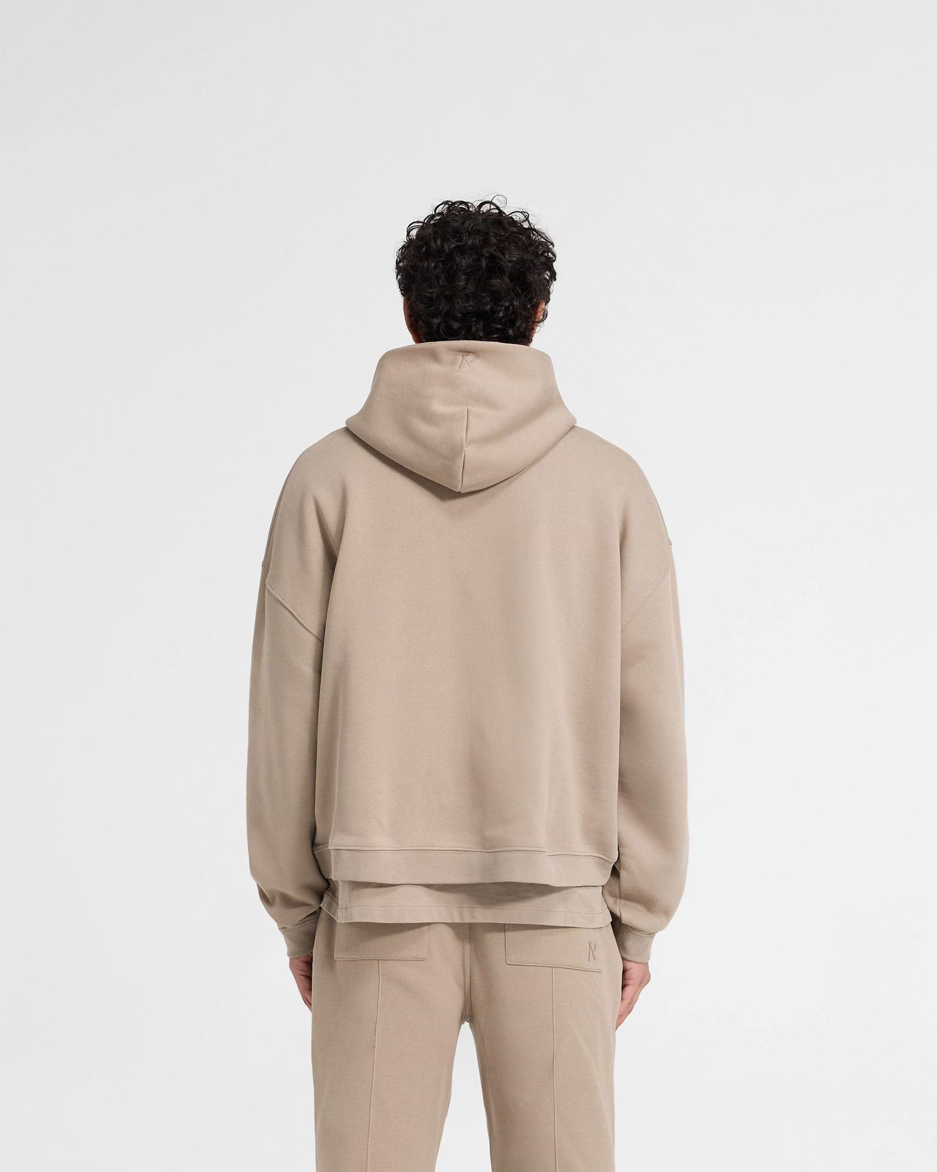 Man wearing an oversized dune hoodie on a plain white background | Initial Boxy Hoodie in Dune | Boxy fit hoodie with cropped body and dropped shoulders | Initial Hoodies | Represent Clo