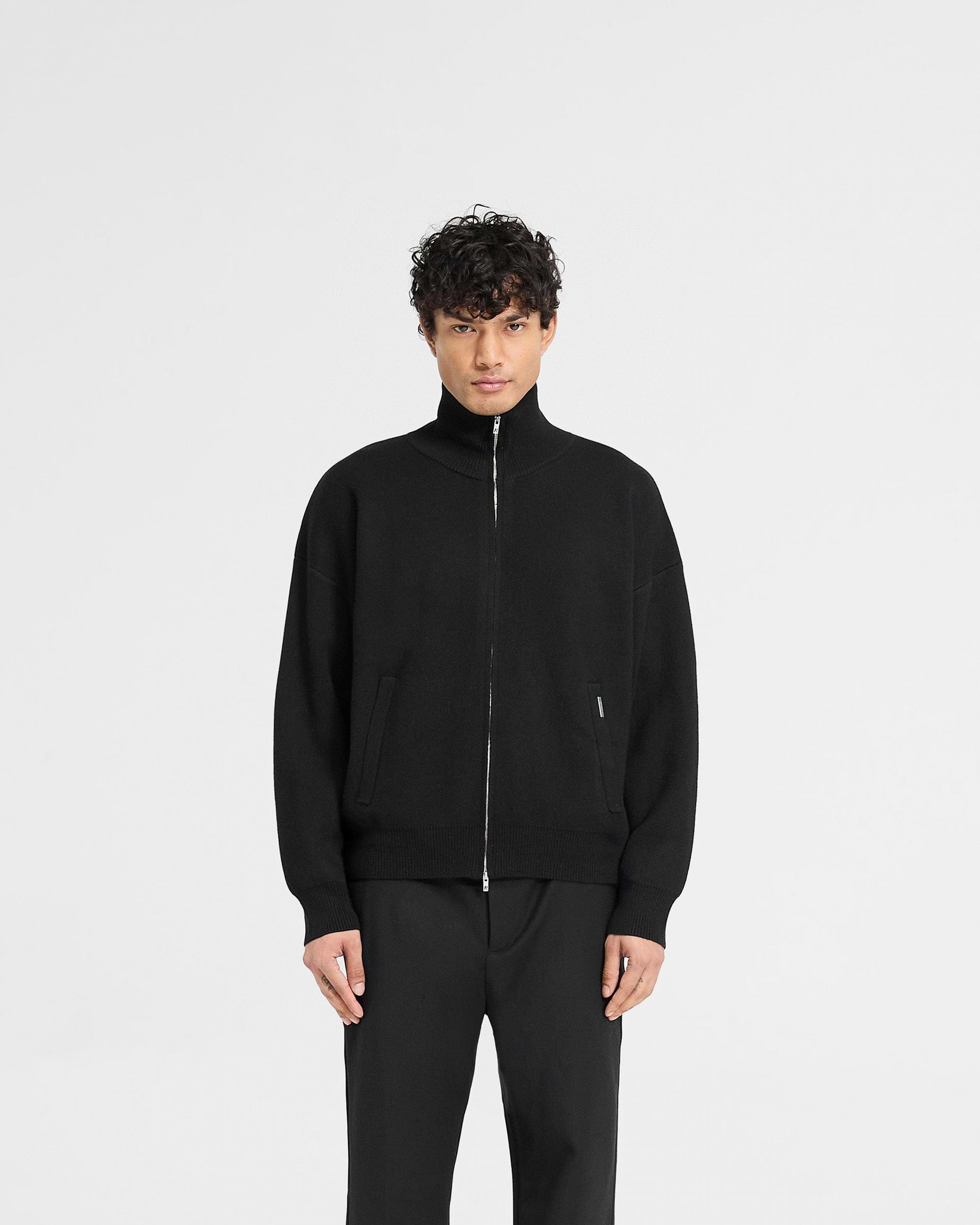 Man wearing a black knitted track jacket on a plain white background | Initial Knitted Track Jacket in Jet Black | Oversized track jacket with zip closure | Initial Knitwear | Represent Clo