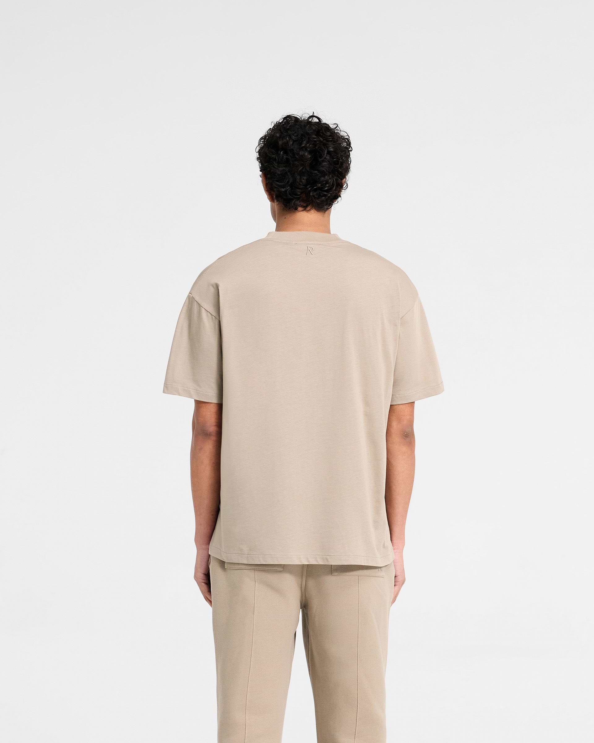 Man wearing an oversized dune t-shirt on a plain white background | Initial Oversized T-Shirt in Dune | Oversized t-shirt with embroidered details | Initial T-Shirts | Represent Clo