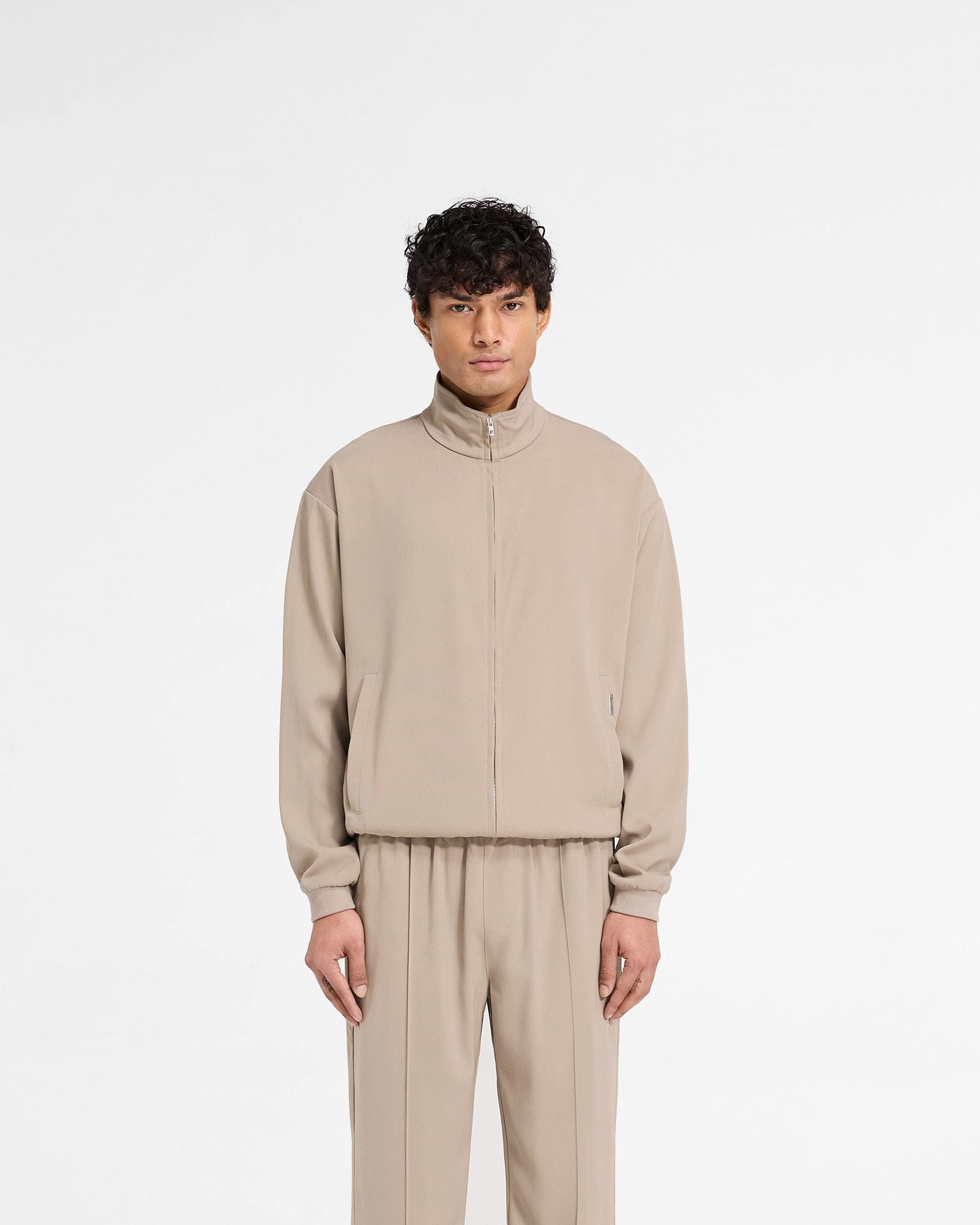 Man wearing a mens beige track jacket on a plain white background | Initial Track Jacket in Dune | Track jacket with oversized fit and RiRi zip | Initial Outerwear | Represent Clo