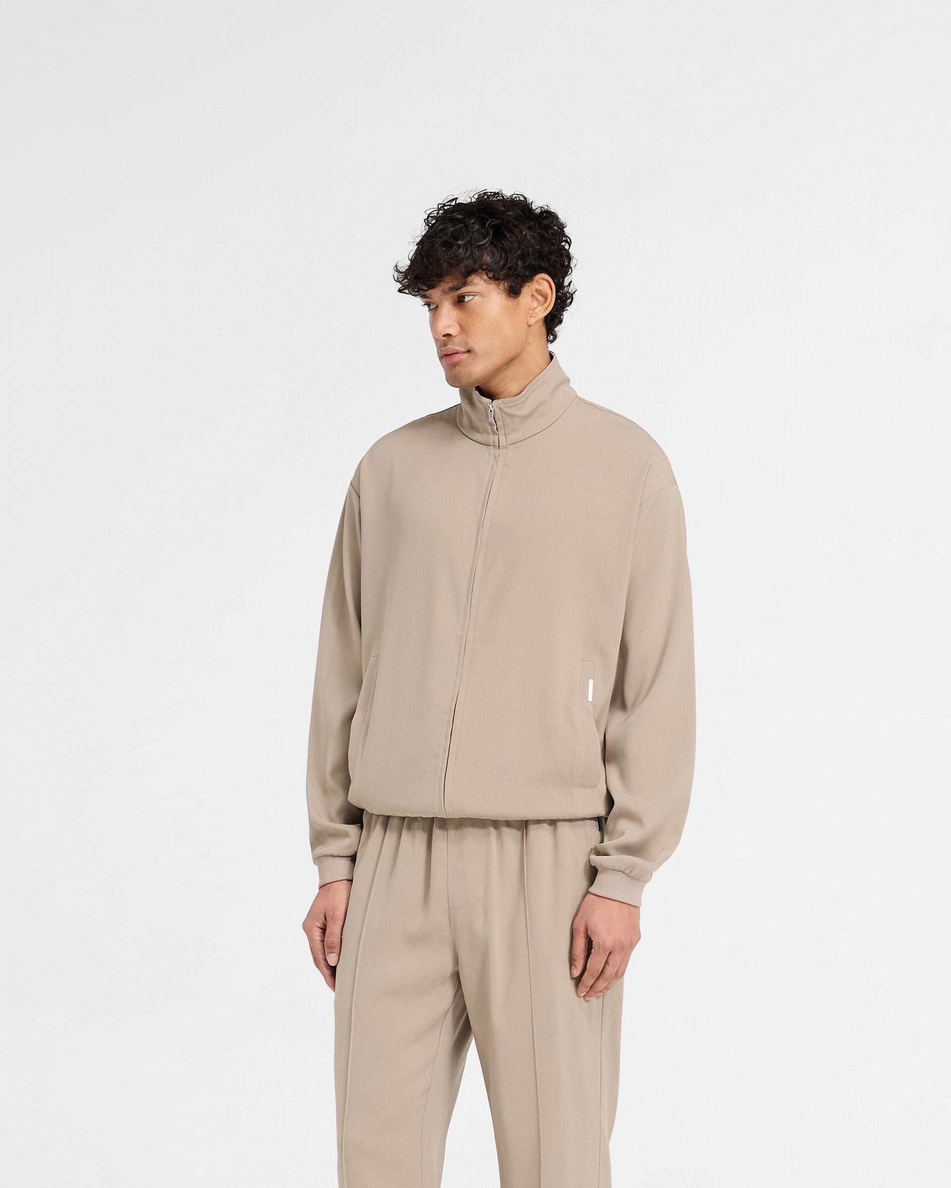 Man wearing a beige track jacket on a plain white background | Initial Track Jacket in Dune | Drapey fabric oversized track jacket with welt pockets and M4 RiRi zip | Initial Outerwear | Represent Clo