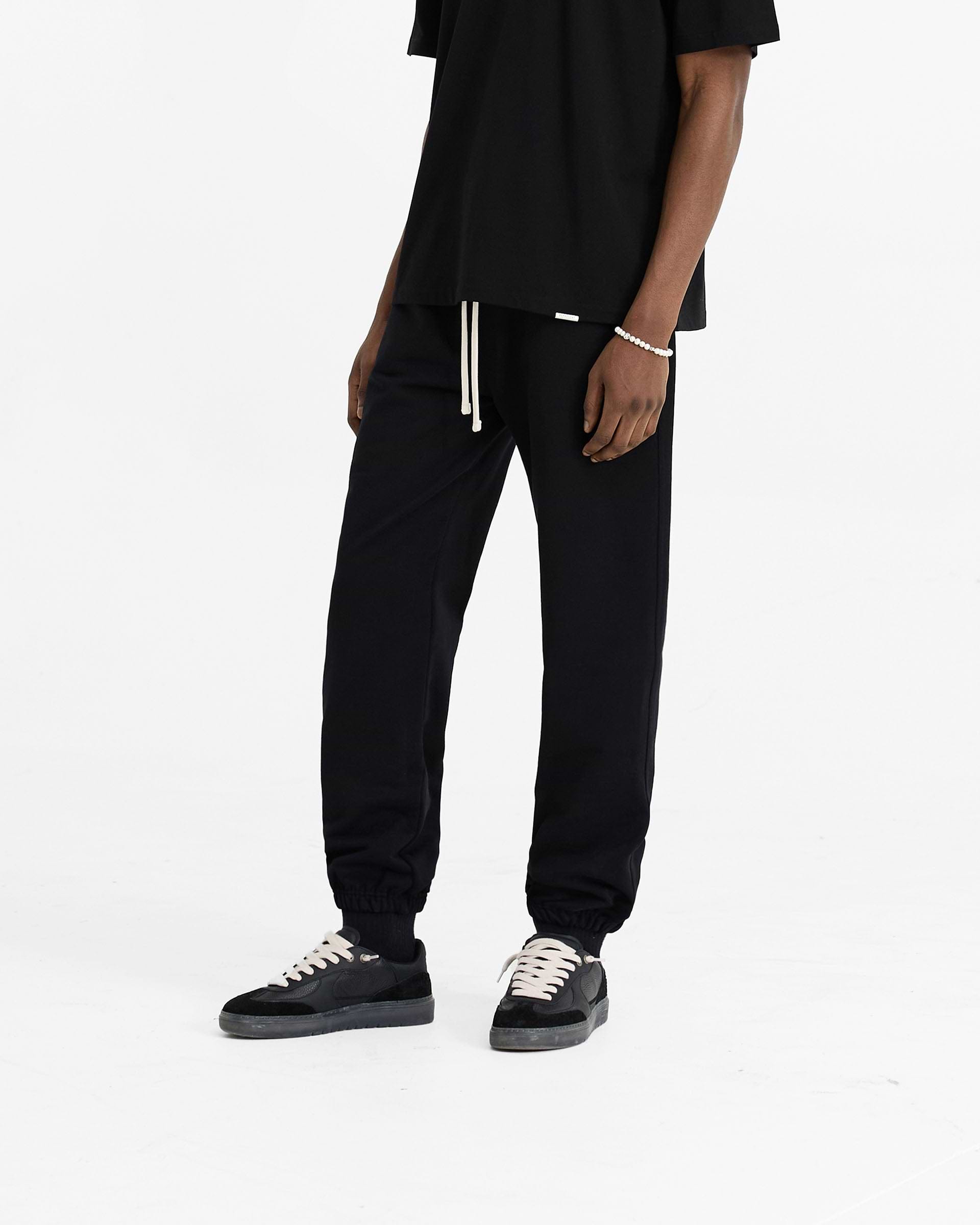 Man wearing black sweatpants on a plain white background | Initial Cuffed Sweatpant in Black | Sweatpants with elasticated waistband and cuffed hems | Initial Pants | Represent Clo