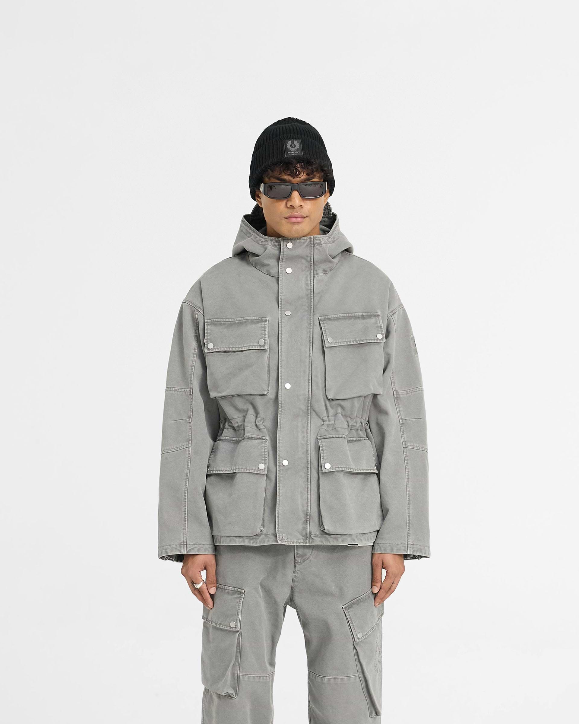 Man wearing a grey field jacket on a plain white background | Represent X Belstaff Enduro Jacket Shadow | Oversized cotton field jacket with adjustable hood and pockets | Exclusives Outerwear | Represent Clo