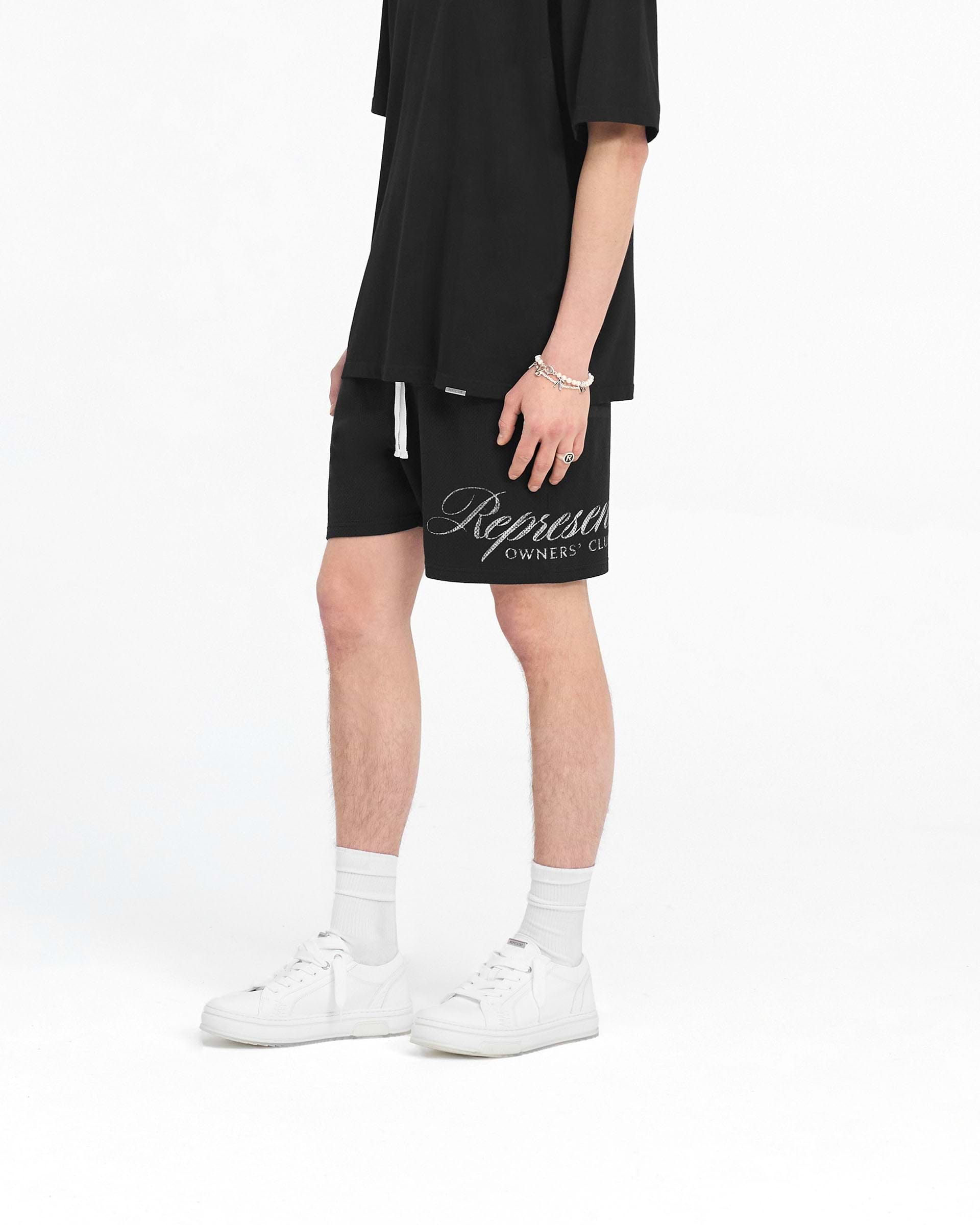 Man wearing black mesh shorts on a plain white background | Represent Owners Club Script Mesh Shorts in Black | Shorts with elasticated waistband and zip pockets | Owners Club Shorts | Represent Clo