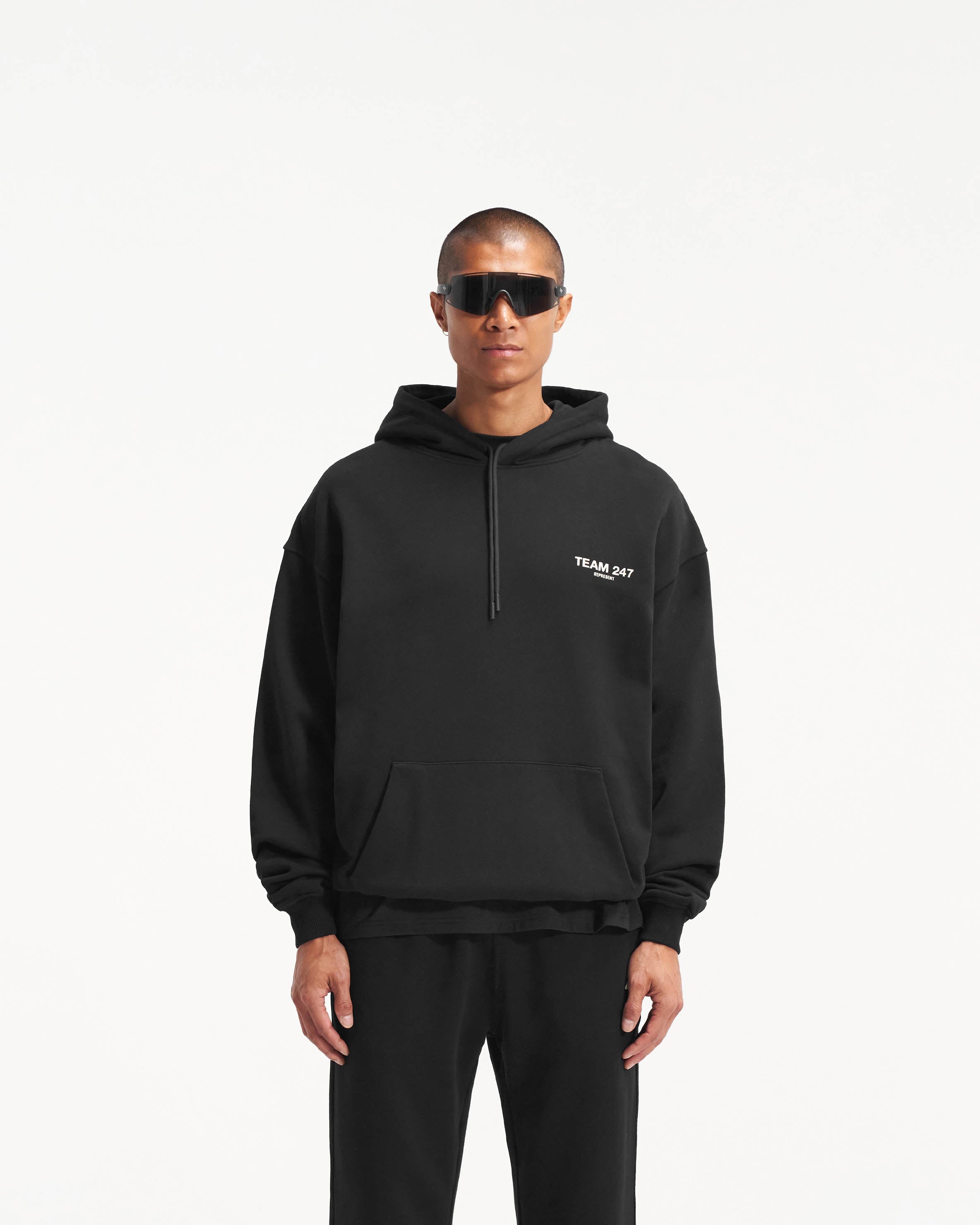Man wearing a black oversized hoodie on a plain white background | Team 247 Oversized Hoodie in Black | Oversized hoodie with ribbed cuffs and Team 247 print | 247 Hoodies | Represent Clo